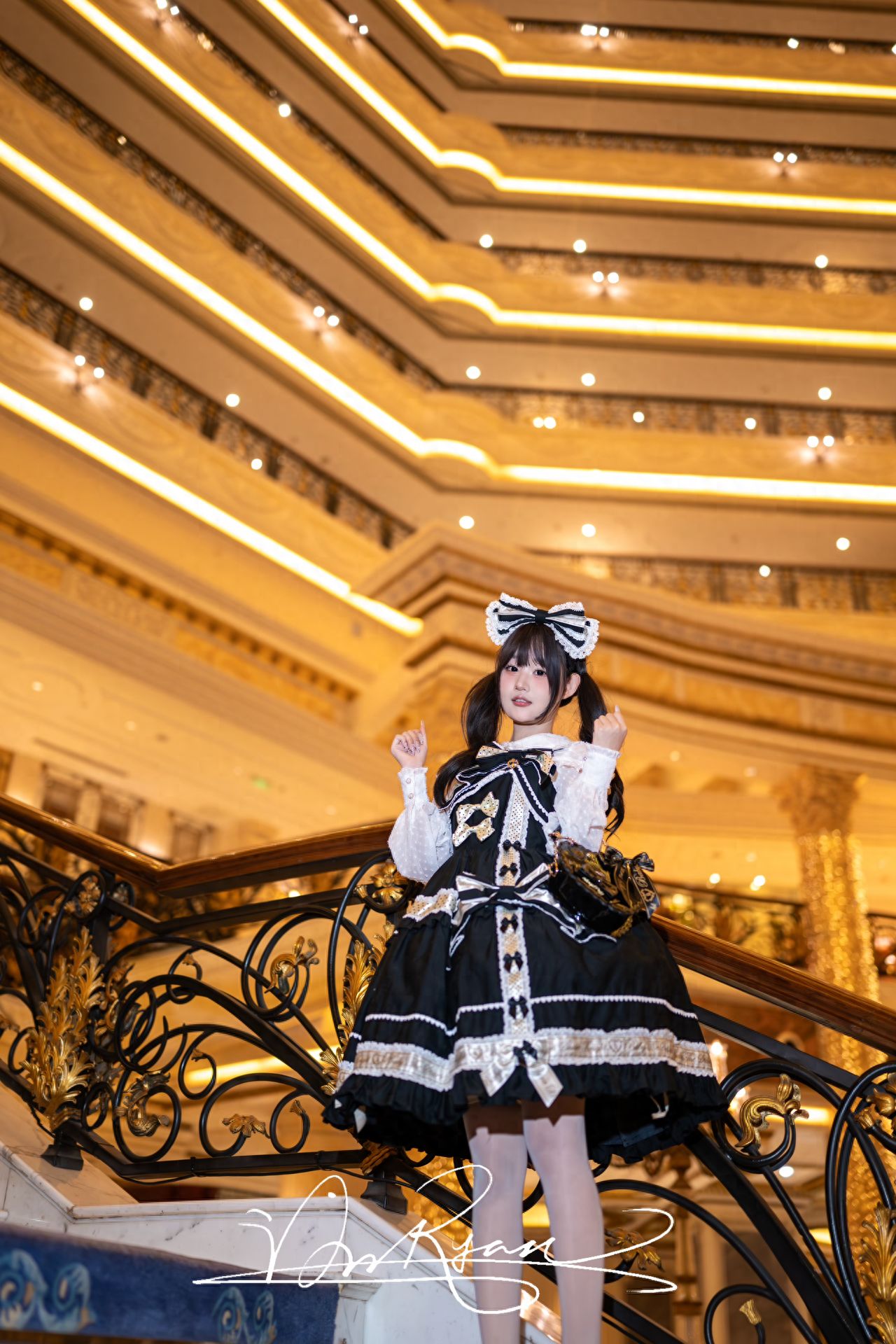 Use a low-angle shot with a centered composition, placing the subject slightly to the left of the center to highlight the main subject and the ornate multi-layered golden ceiling above. Utilize the curve of the stair railing and the extended lighting lines of the ceiling to naturally guide the viewer's gaze towards the subject. The frame should include the golden ceiling background and the stair railing foreground to enhance the sense of depth in the image. The model should raise both hands to make a cute gesture, adding a lively, interactive feel to the photo. Clothing suggestion: Consider wearing Lolita attire, which is very cute and photogenic.