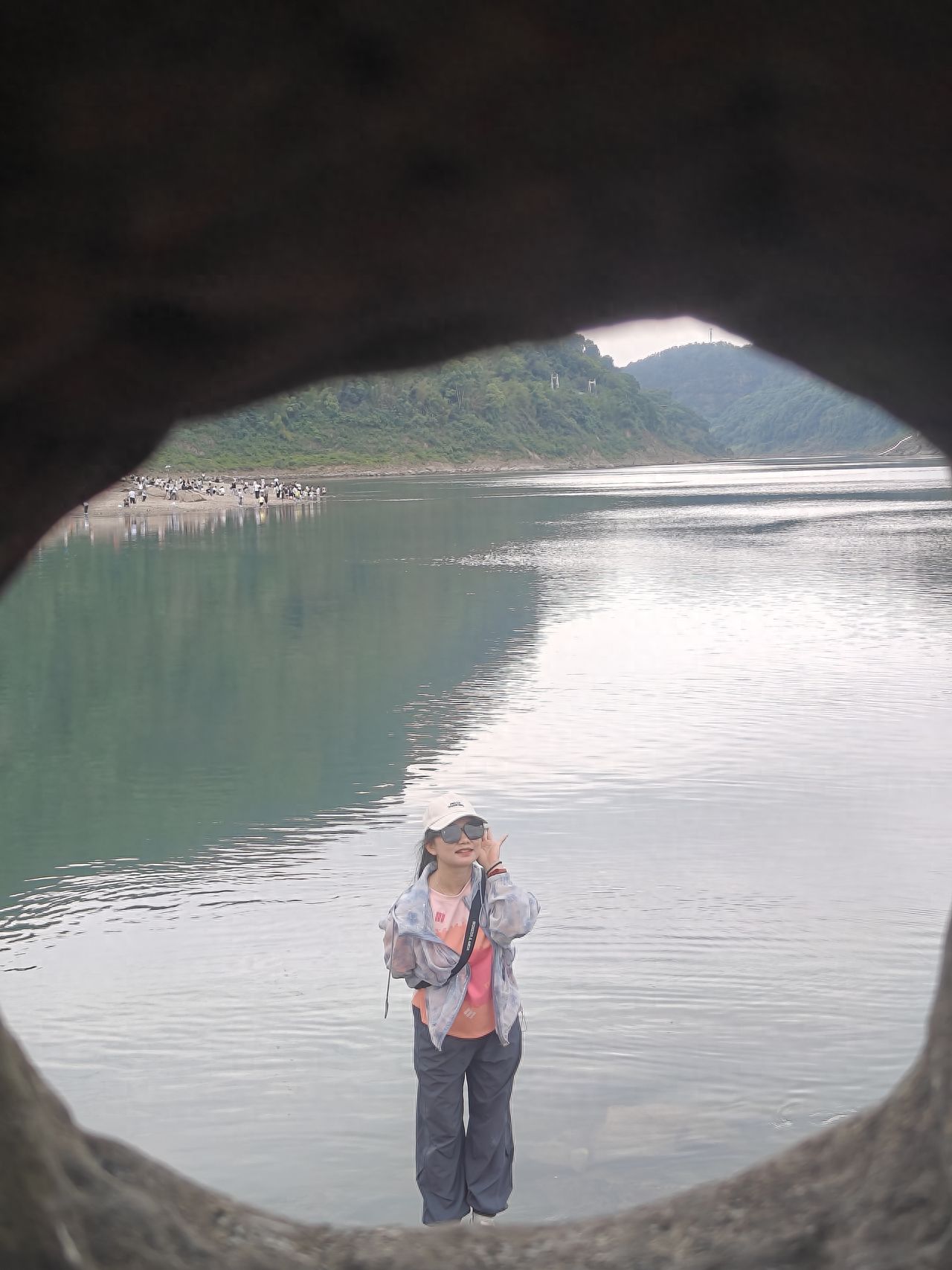 Use the cave entrance as a foreground frame for composition, with the model standing by the water, centrally located in the frame, and the photographer adopting a level shooting angle. The shot includes the foreground cave entrance rocks, the midground characters and water surface, the background mountains, and the people on the shore, enhancing the sense of depth. It is suggested to use a standard focal length/phone 2x for shooting.