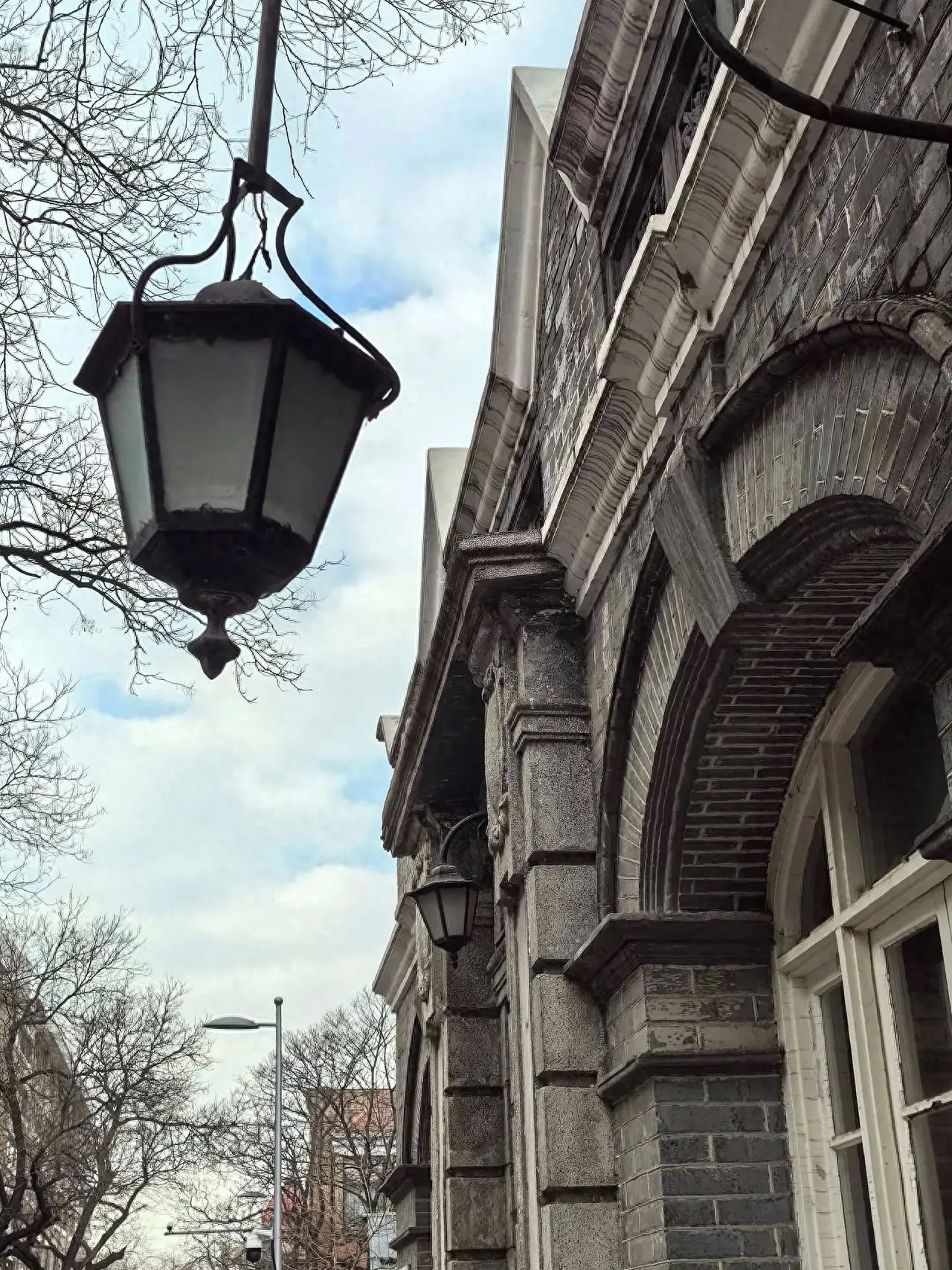 The photographer stands on the side of the entrance door lamp, using a slightly upward angle for the shot. The lamp is placed in the upper left corner of the frame, with branches, blue sky, and white clouds as the background, and the former French Post Office building on the right side. Additional experience: Not open to the public for now.