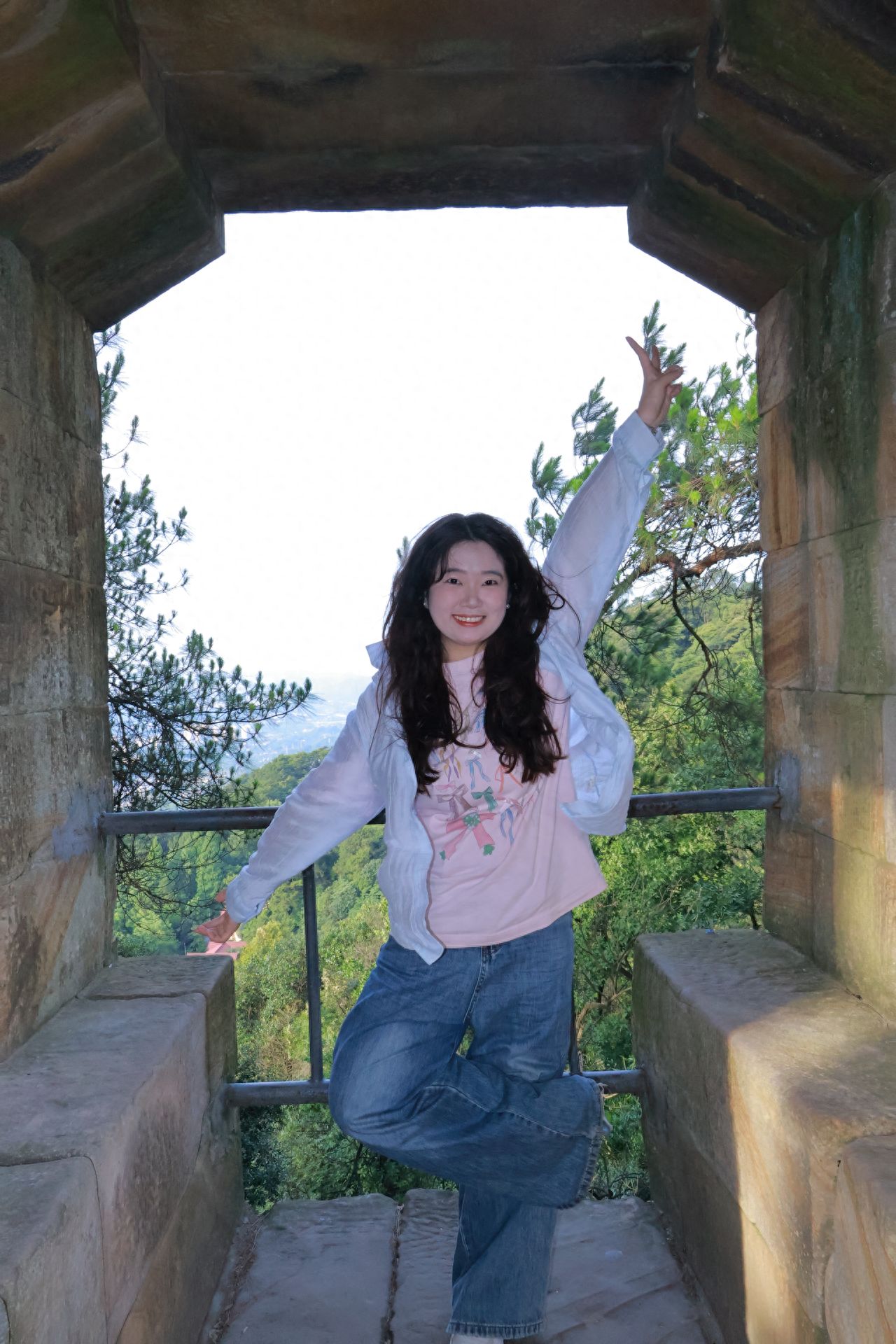 Utilize the stone door frame as a compositional framework, with the model standing in the middle of the passage, centrally located in the frame, and the photographer shooting from the inside of the door frame at eye level. The scene should include the door frame structure, background green mountains and trees, and railings to enhance the sense of depth. The model can either place their hands on their hips to show confidence or lean casually on the railing, with postures that are relaxed and natural. Use a wide-angle lens or the 1x lens on a smartphone.

Photography poses:
1. The model adopts a hands-on-hips stance, with both hands on their waist, showcasing a confident and relaxed state that highlights body lines, conveying a sense of ease and a touch of vigor;
2. The model naturally drapes their arms over the railing, maintaining a relaxed and casual posture, creating a cozy and spontaneous atmosphere, as if they are enjoying the surroundings at leisure, providing a visually comfortable and relaxed feeling. Both poses fit the outdoor scene, displaying different emotional states.

Travel tips: From 5-7 PM, as the sun sets, the remaining light casts a warm golden hue on the body and the surrounding rocks and trees, making it an excellent time to capture atmospheric photos.