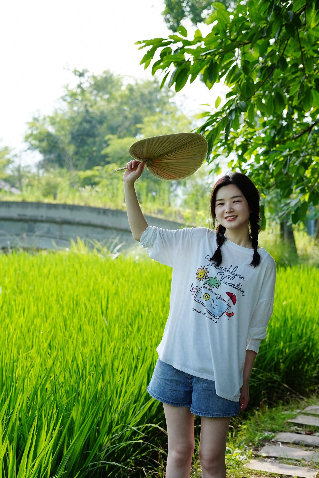 Using the rule of thirds composition, place the model at the right third of the frame, with the photographer shooting from a flat angle to match the natural perspective; include a large expanse of green rice fields in the background, and retain leaves in the right foreground to add depth. 1. Pose suggestion: The model holds a straw hat with one hand above their head, standing naturally and smiling. 2. Wardrobe suggestion: It is recommended to wear a white tank top paired with light-colored denim shorts.