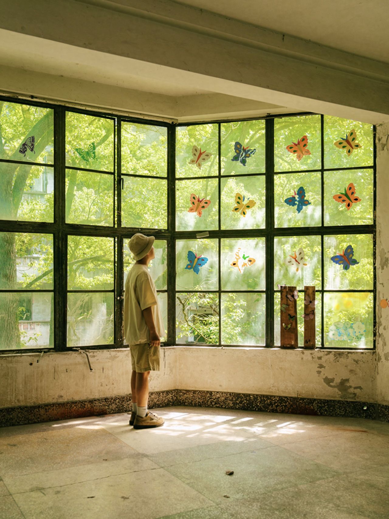 Use a centered composition with the subject at the center of the frame and a flat shooting angle. The model stands in front of the window, looking up at the glass with butterfly stickers, with a natural posture. Include a large area of the window, green trees outside the window, and the light and shadow on the ground to enhance the sense of space. Additional tips: The abandoned kindergarten's gates are locked, so enter by climbing over the wall, and pay attention to safety.