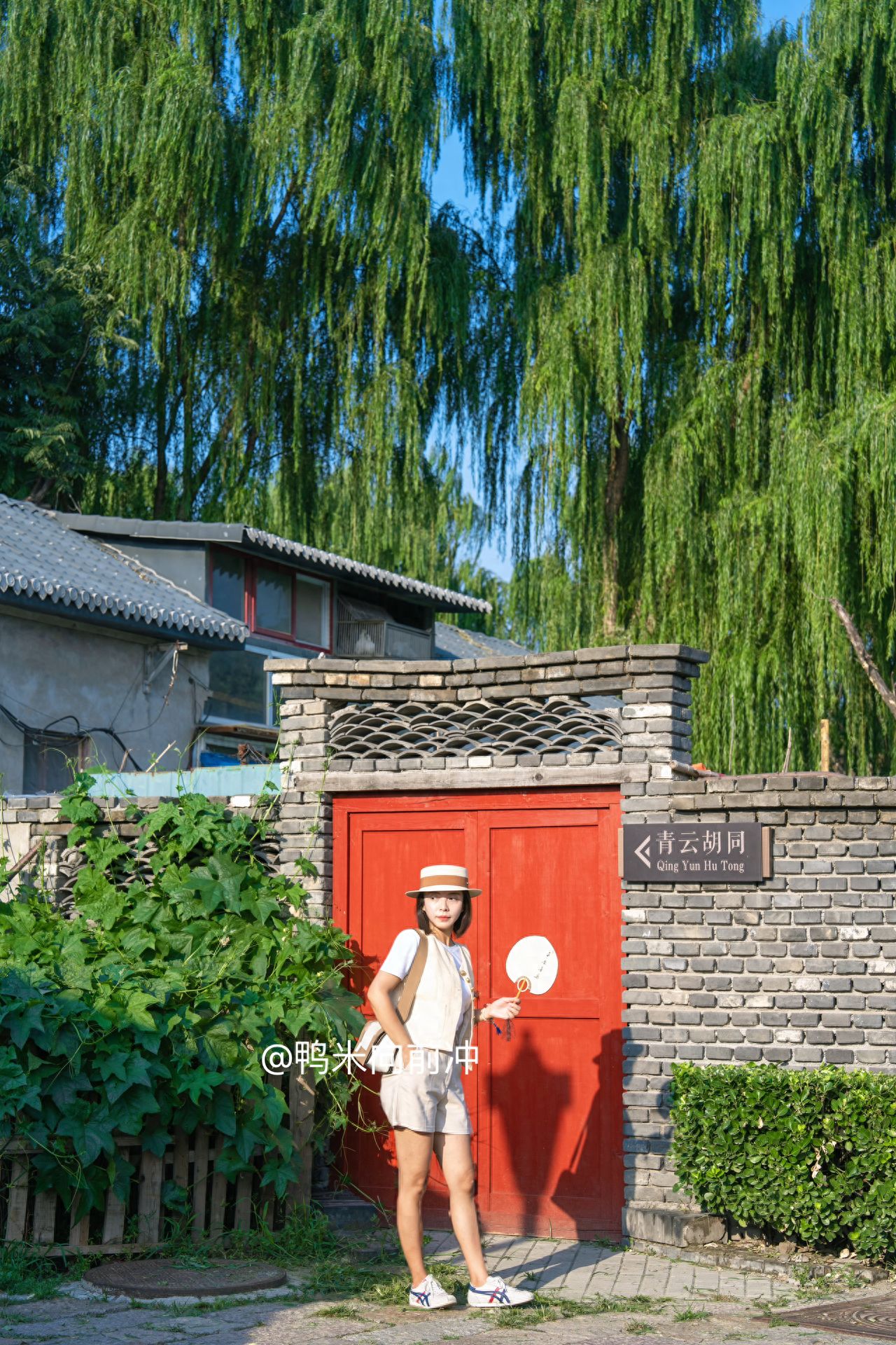 Using the rule of thirds, the model is positioned at the bottom third of the frame, with the red door at the center to balance the visual composition. The photographer slightly tilts the camera upwards, capturing the red door, brick wall, greenery on the left, and the weeping willows in the background to enhance the depth of the scene.

Pose for the photo: The model stands to the left of the red door, holding a folding fan and standing naturally with a relaxed posture.