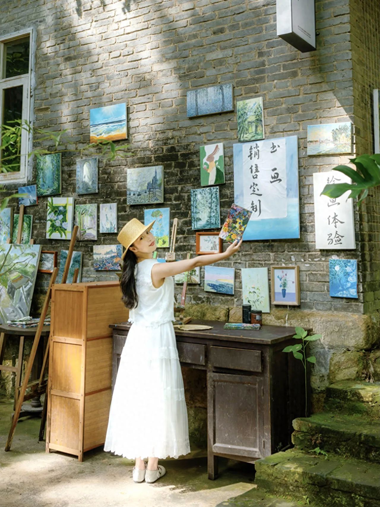 The model is positioned slightly to the left in the center of the frame, holding a painting and gesturing towards the wall, turning her head and smiling to enhance the interaction with the environment. The photographer takes a frontal shot, capturing the full wall of decorative paintings, an old wooden table, and the paint tools on the table, preserving the scene's details. 
Travel tips: The admission fee is free.