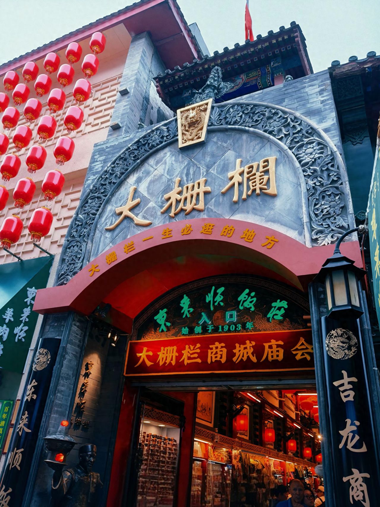 The photographer stands below the plaque and takes a photo looking up to highlight the towering feel of the architecture. Then, use a centered composition with the "Dashilar" plaque in the center of the frame to ensure the text is clear and complete. The scene includes the string of red lanterns on the left, the storefront on the right, and the carved roof above, showcasing the atmosphere of the ancient street. It is recommended to use a wide-angle/phone 1x lens to capture more environmental details.