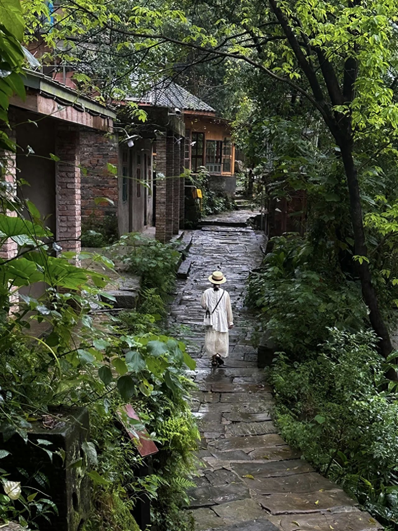 The photographer is positioned at a higher spot behind the ancient village street, capturing the scene from an aerial perspective. The cobblestone road serves as the main guiding line in the frame, with the figure's white attire creating a stark contrast against the surrounding dark green vegetation and quaint red brick buildings. The greenery and architecture on both sides of the street seem to envelop the central cobblestone path and the figure. It is recommended to use a medium-telephoto lens.

Outfit suggestion: A light-colored dress is recommended.
