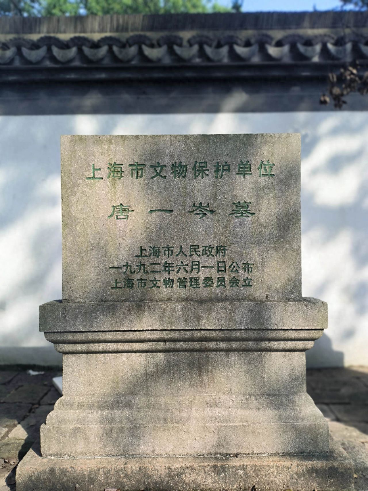 The stone tablet is centrally located in the frame, highlighting the main inscription. The photographer is capturing it in a frontal, flat shot to ensure that the text is clear and complete. The background includes a white wall and tiles on the roof above, while the foreground contains the base of the stone tablet and some of the ground bricks, enhancing the sense of environment and depth.