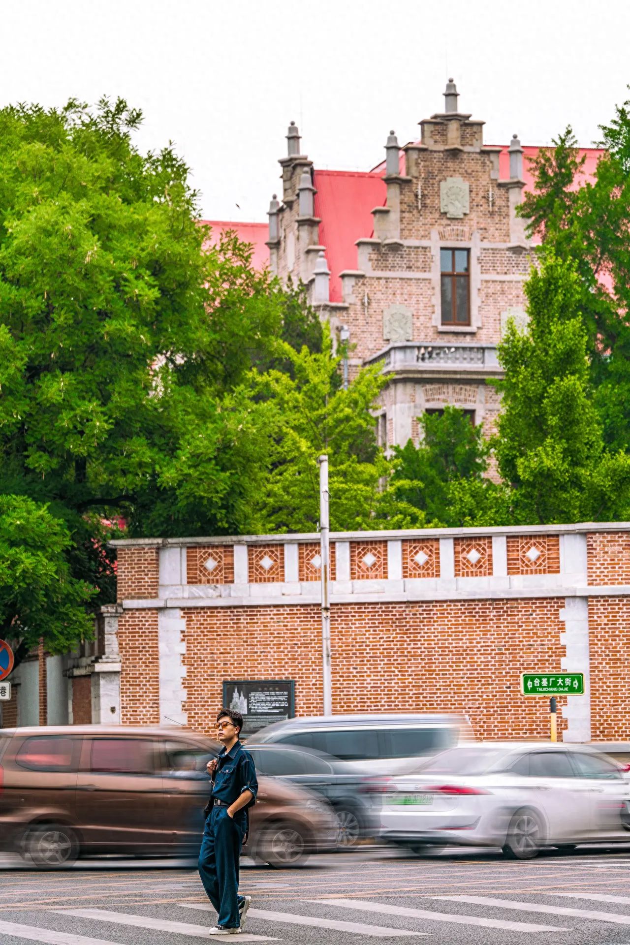 The model stands on the zebra crossing while the photographer captures the scene with a medium telephoto lens, including the model, road, vehicles, European-style buildings, red brick walls, and trees in the frame. The red-roofed European-style building is centrally located to highlight the subject, and the brick wall serves as a background to enhance the color contrast in the image. 

Travel Tips: Shoot on a sunny day for better lighting and more impressive photos.