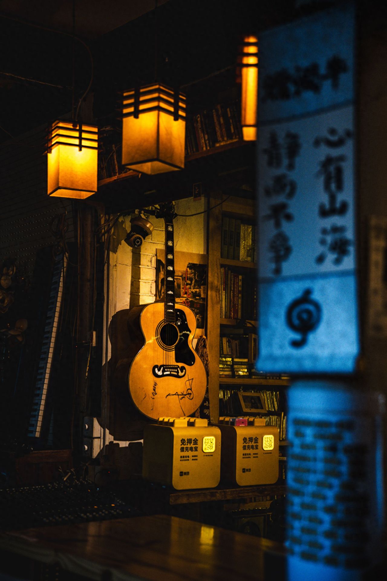 Use the rule of thirds to compose the shot, place the guitar slightly off-center to the left to highlight the subject. Incorporate and blur the text signboard on the right for depth, capture the bookshelf, books, and hanging lights in the background to create an indoor atmosphere. Use a flat angle to ensure the subject is clear and environmental elements are intact.