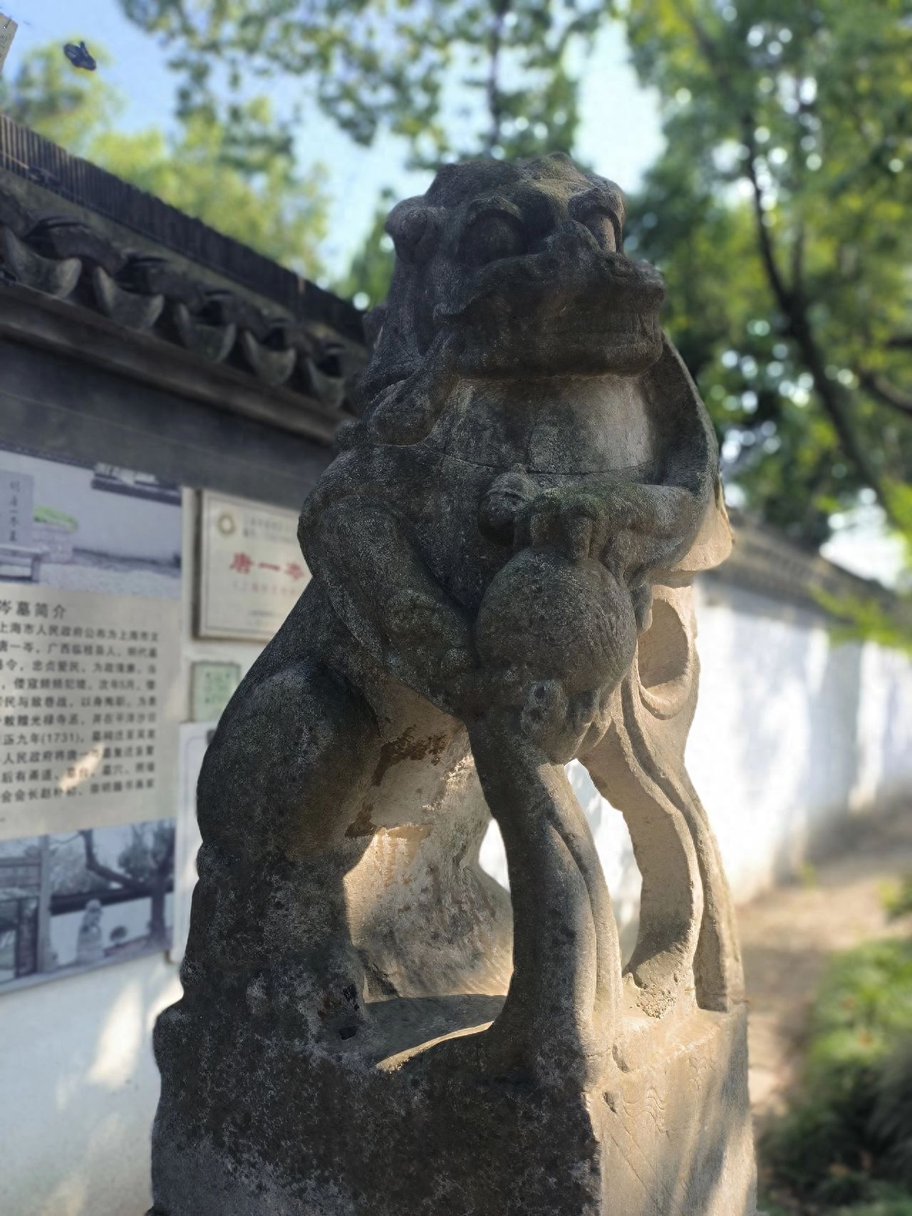 Compose the shot with the stone lion at the center of the frame to highlight the details of the sculpture. The photographer should shoot from a level angle, aligning with the height of the stone lion. Include the roof of the ancient building on the left and trees on the right in the frame, and keep the sky in the background to enhance the atmosphere. It is recommended to use a wide-angle lens to capture both the subject and the surrounding scenery.