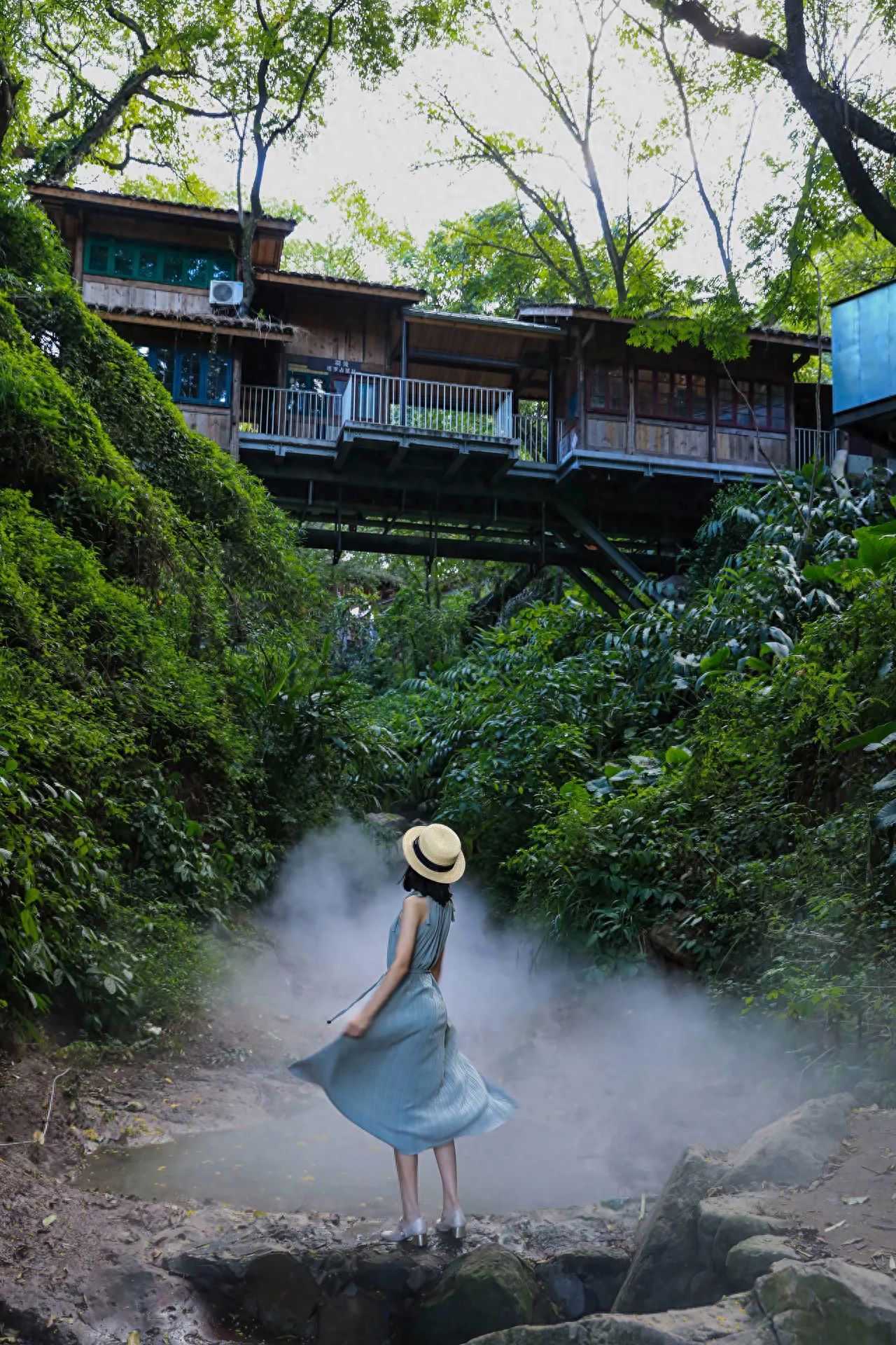 The photographer uses a low-angle shot with the model positioned centrally at the bottom of the frame, incorporating the wooden building at the top third to enhance the sense of depth. The composition includes the wooden building with a balcony in the background, the foreground stream rocks and mist, and retains green vegetation around. The model faces away from the camera with her skirt naturally flowing and interacting with the mist to add a sense of movement. A standard focal length is used for the shot. Additional advice: it is recommended to take sun protection measures.