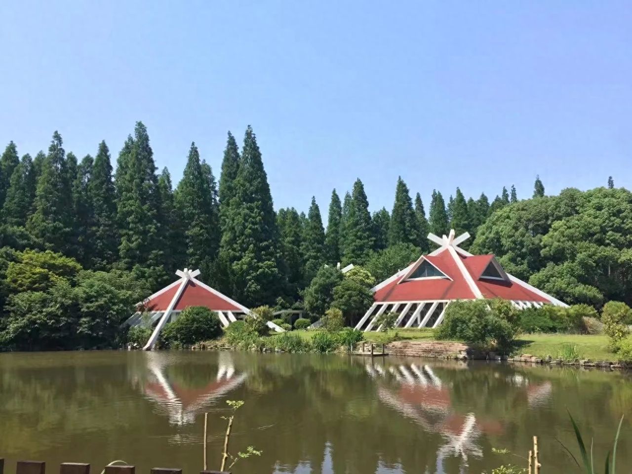 Shoot from a low angle with the camera pointing upwards, using the water surface as the foreground to capture the complete reflection of the building in the lake, creating a reflection composition that enhances the sense of balance in the image. The frame should include the two red-roofed main buildings, green trees by the lake, and the blue sky in the background, blending nature with architecture. It is recommended to use a standard focal length to ensure the clarity of architectural details and reflections, and to achieve a well-defined layering in the image.