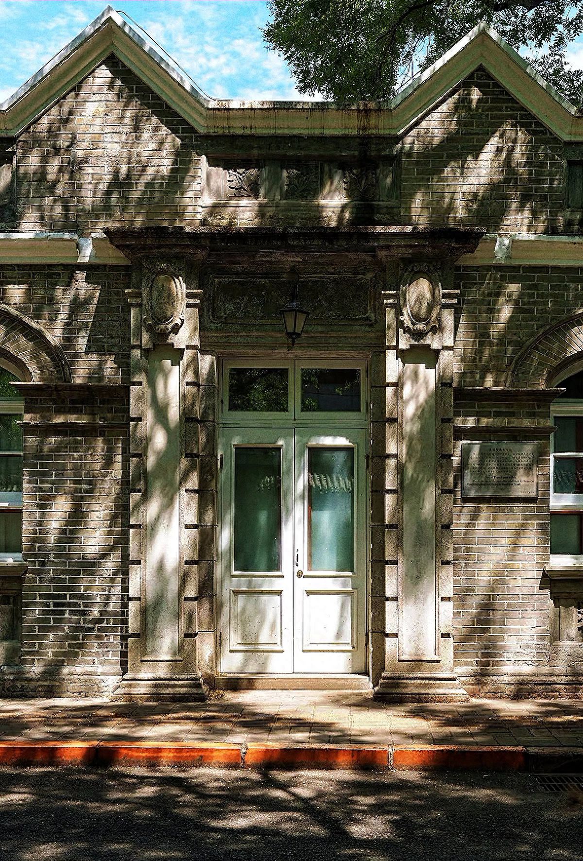 The composition is symmetrical, with the entrance of the building at the center of the frame, and the brick walls, decorations, and arched windows are symmetrically distributed on both sides, highlighting a sense of solemn balance. The photographer uses a low-angle shot to capture the entirety of the entrance, the triangular roof at the top, and the detailed carvings. The frame includes the stone steps in front of the door, the light and shadow on the walls, and retains some sky and tree branches at the top to enhance the sense of depth in the image. It is recommended to use a medium telephoto lens to compress the space and highlight the texture and structural details of the building. Travel tips: no admission fee required.