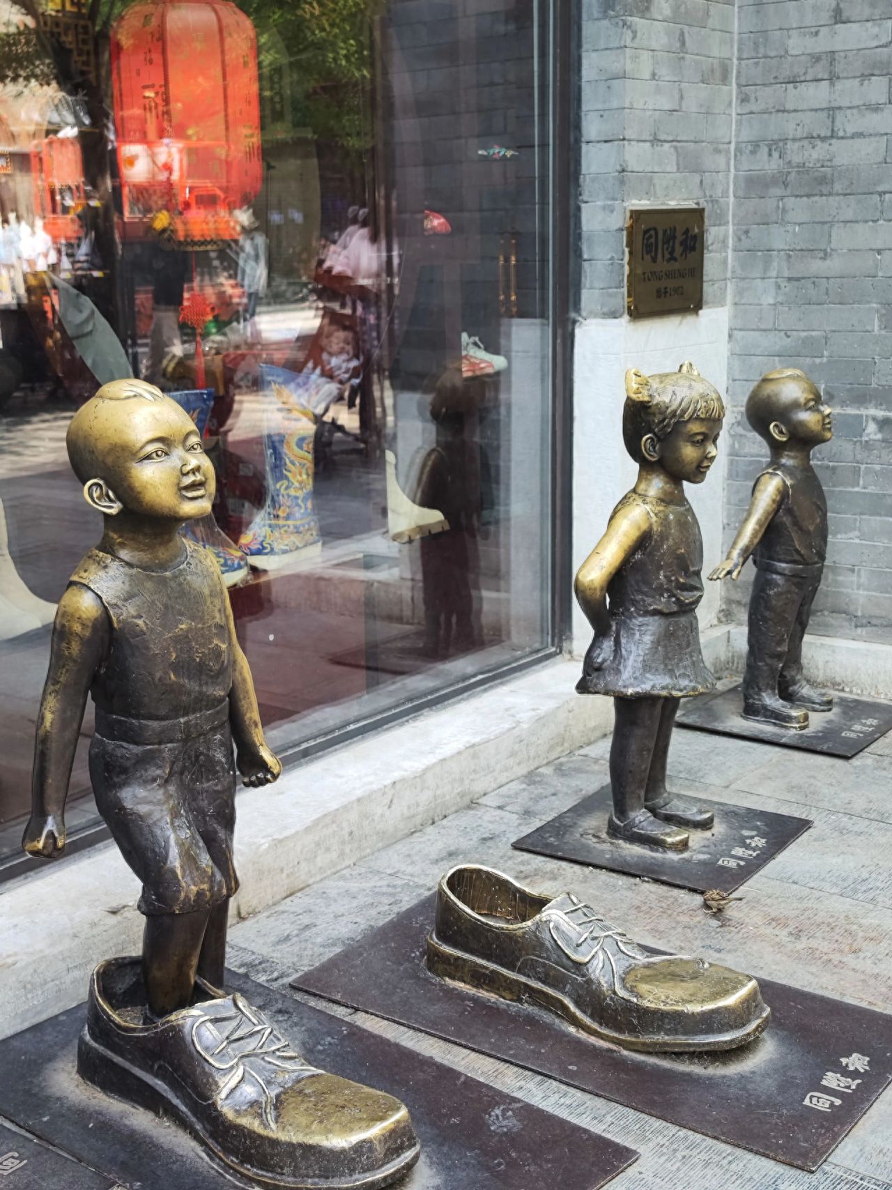 Capture the shot from a high angle, placing the children's sculptures and shoe sculptures in the center of the frame to highlight the main group. Include the shop's glass windows and brick walls in the background, and keep the stone slabs on the ground in the foreground to enhance the sense of depth in the environment.