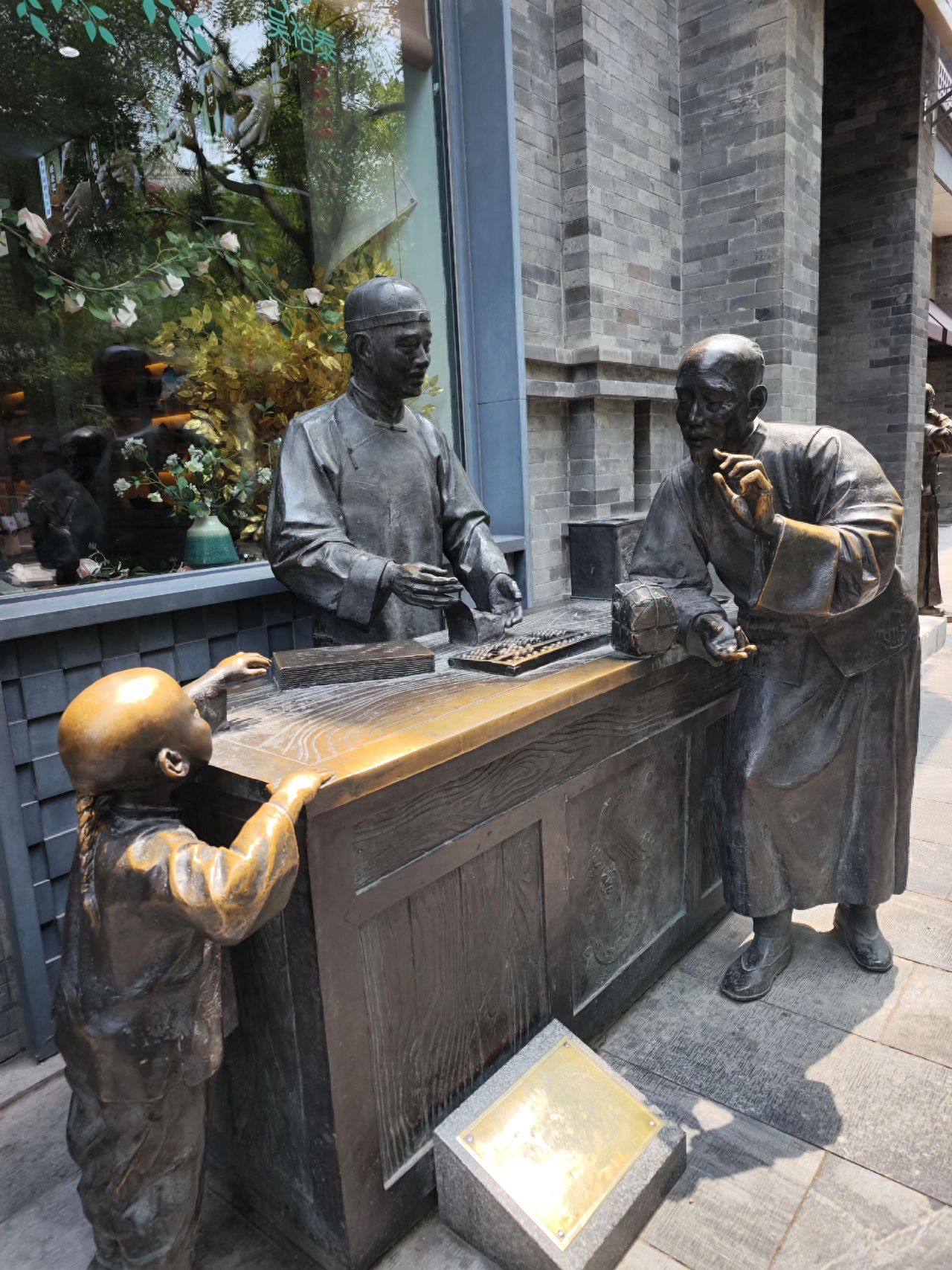 Adopting a bird's-eye view, the photographer stands at a slightly elevated position to place the three bronze statues and the entire counter in the center of the frame, highlighting the interactive scene. The background includes the teahouse's glass window (with green plants and decorations) and the gray brick wall, showcasing the ambient atmosphere.