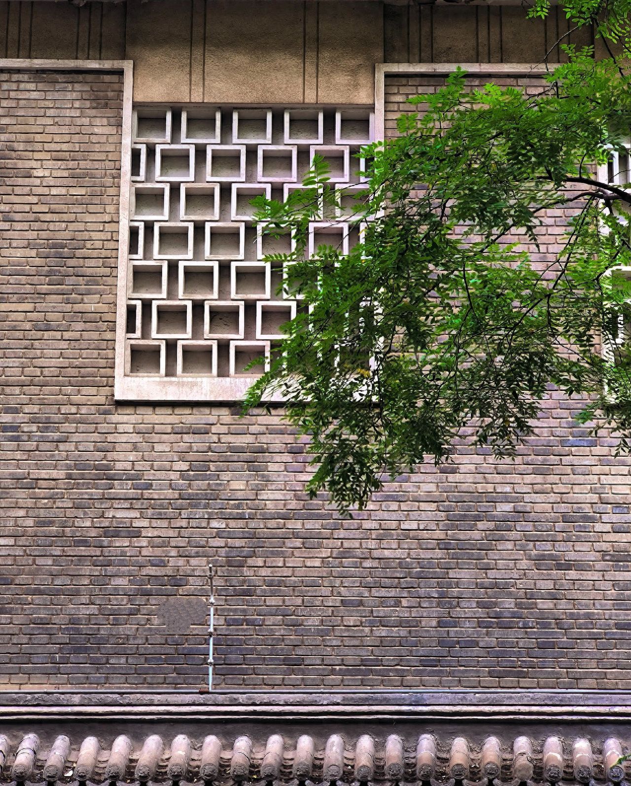 The photographer uses a low angle shot, capturing from the bottom up. They employ frame composition, placing the square grid window at the center of the frame as the main subject, and incorporating natural branches on the right to add depth. The framing includes the full view of the brick wall, the window, and the tiles below, preserving environmental details. A telephoto lens is recommended to emphasize the contrast between the main subject and natural elements.