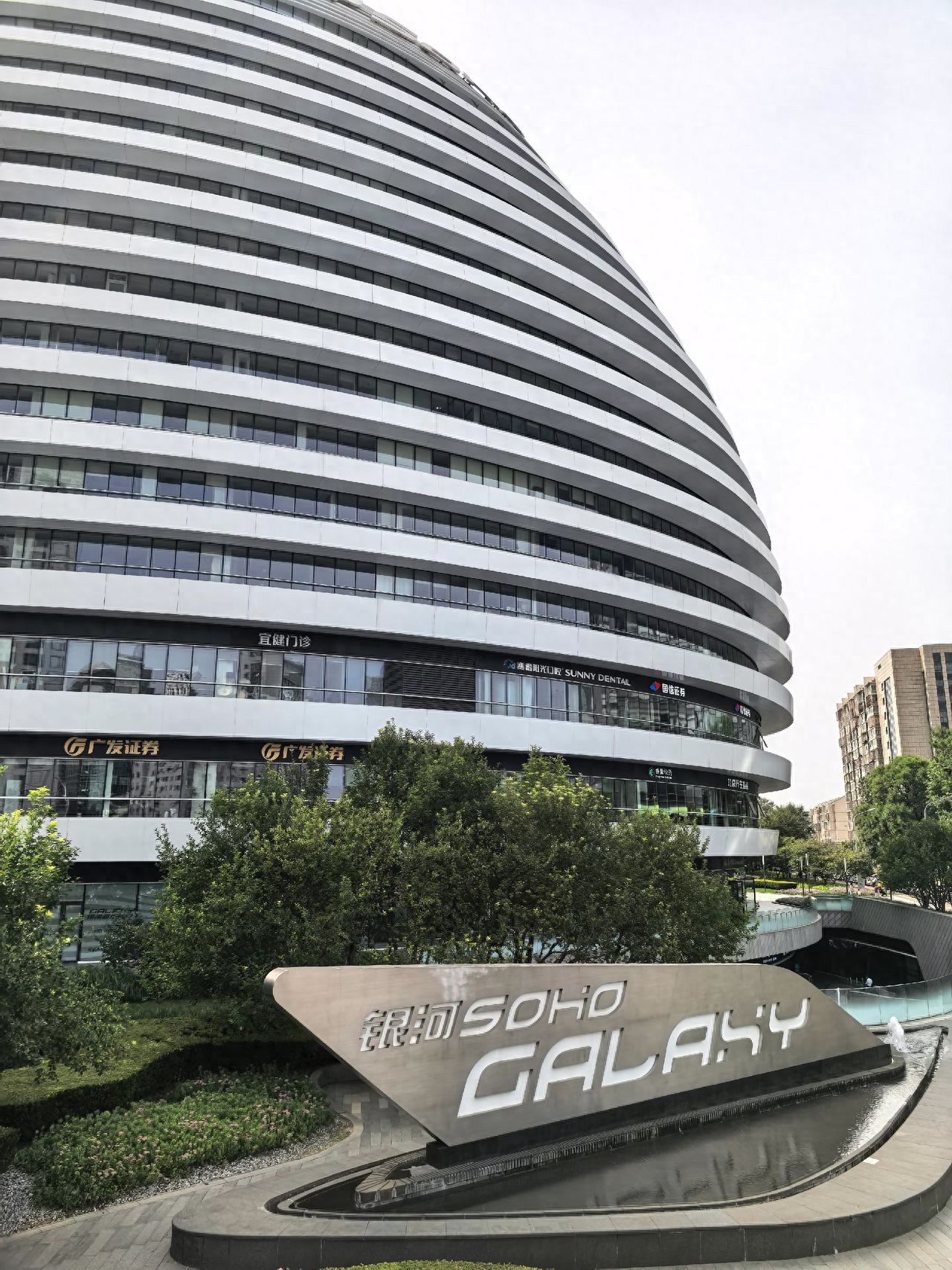 The photographer employs a low-angle shot to emphasize the towering height and curved lines of the building, utilizing diagonal composition with horizontal stripes extending from the bottom left to the top right to guide the viewer's gaze. The foreground includes a sign for Galaxy SOHO, and the background encompasses the main structure of the building, enhancing scene recognition.

Travel Tips: Admission is free.