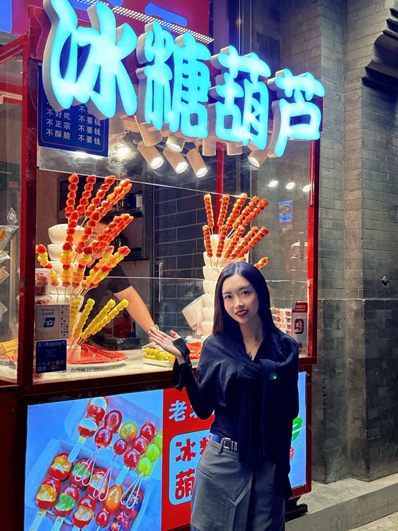 The model stands in front of a candied hawthorn stand, positioned at one-third of the frame from the right, using a flat shooting angle. The composition is arranged according to the rule of thirds, with a blue neon sign and displayed candied hawthorn skewers included on the left, and the model's right hand raised naturally on the right, enhancing the liveliness of the image. It is recommended to use a standard focal length to ensure clarity of the subject and background details.