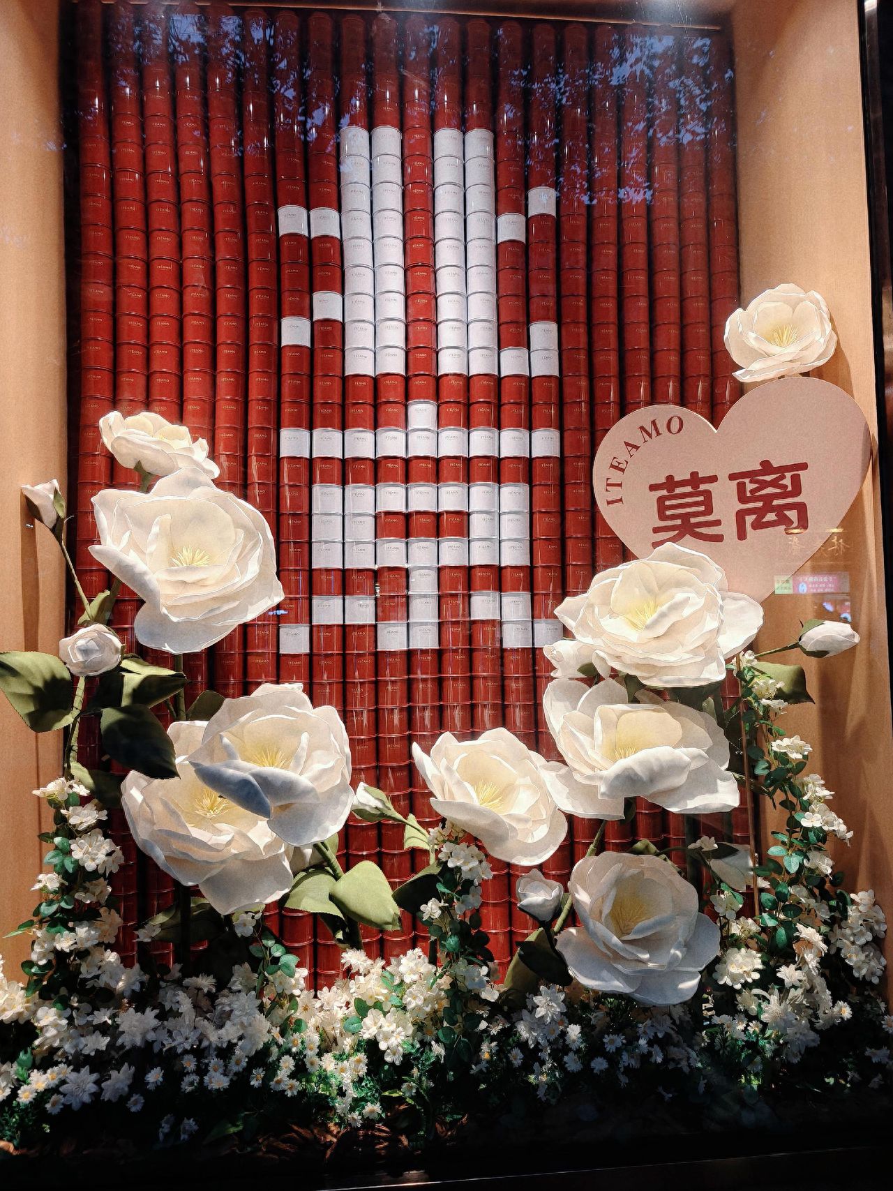 Compose with a foreground and background, using white roses as the visual focus in the foreground, and a symmetrical arrangement of red and white cans in the middle ground to enhance the sense of depth. Ensure clarity of elements in the window display, with the foreground roses, middle ground cans, and the heart-shaped decoration in the upper right corner. Use a standard lens to capture the image, avoiding reflections from the glass that could disrupt the cleanliness of the shot.

Additional tips: Avoid peak traffic times and travel during off-peak hours.