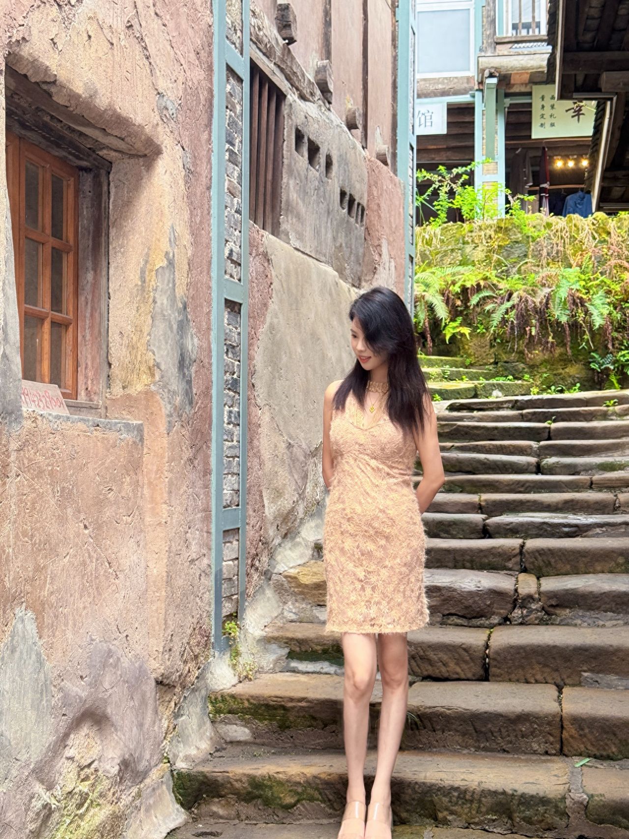 The model is positioned on the right side of the frame, with the scenery including the mottled vintage wall with wooden windows and blue frames on the left, and the stone steps extending upwards with green plants on the right, enhancing the sense of depth in the environment. The model stands with hands behind the back, looking down with a natural posture that blends into the vintage atmosphere.

Additional Tips: It is recommended to wear comfortable shoes, as there is a lot of walking within the scenic area.
