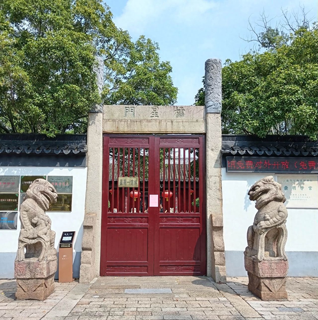 Stand slightly lower in front of the main gate and use a low angle with a tilt upwards to emphasize the solemnity of the gate. Place the gate in the center of the frame with the stone lions symmetrically distributed on both sides to enhance the sense of balance. Include the sky above, green trees on either side, and the stone path leading to the gate to highlight the main subjects of the gate, the plaque, and the stone lions.